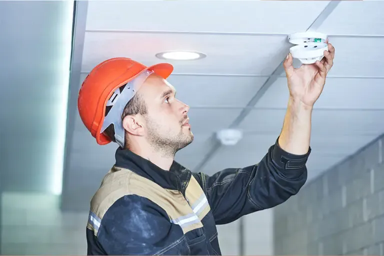 Engineer in a hard hat reaching up to a ceiling-mounted fire detector